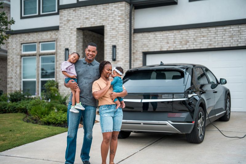 A cheerful multiracial family gathers by their parked electric vehicle, transferring shopping bags in front of their solar-paneled residence, embodying sustainable living.
