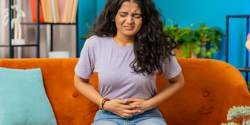 Young woman sitting on couch, holding stomach in pain.