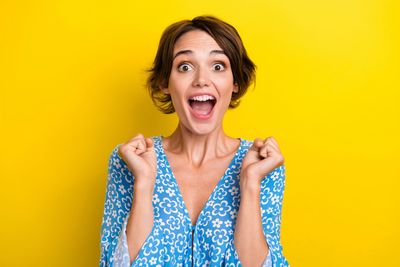 Excited woman in blue dress expressing joy with clenched fists against yellow background.