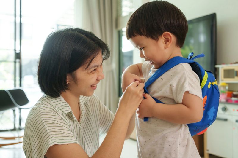 Smiling Asian mother adjusts blue backpack on happy young son. Close-up in living room. Child laughs while mother ensures straps are secure. Parent-child bonding, joy, and morning routine.
