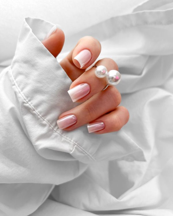 Hand with glossy pink French manicure and pearl ring on white fabric.