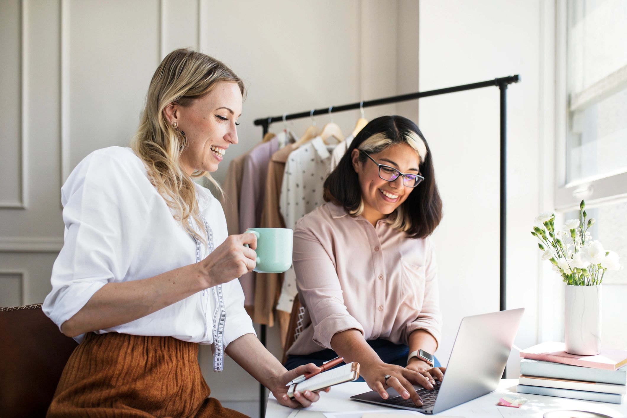 Two women collaborating happily on a laptop in a bright, stylish workspace.
