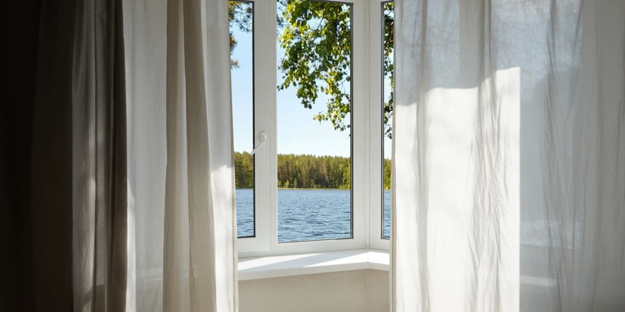 View of lake and trees through open window with sheer curtains.