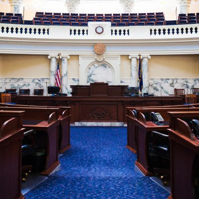 Empty legislative chamber with wood desks and blue carpet.