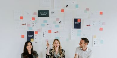 Three people standing next to a wall that has charts and sticky notes on it