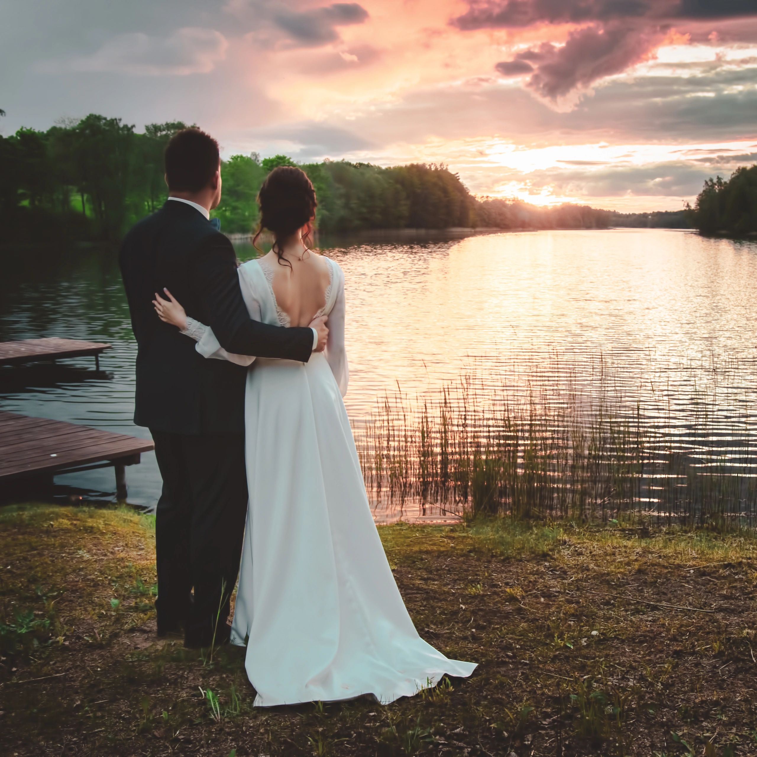 A couple in wedding attire embraces by a lakeside at sunset.
