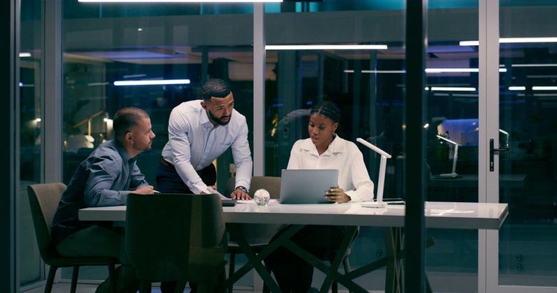 Overtime, male leader and team in office with laptop, discussion and brainstorming at night. Late work, diversity and collaboration in boardroom for research, advice and talking in modern workplace