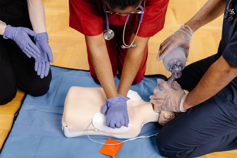 Three people are practicing CPR on a training mannequin. One person is performing chest compressions while another is using a bag valve mask to give breaths.