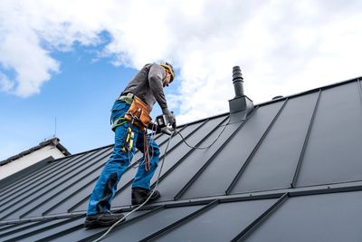 A worker in safety gear drills on a metal roof under a blue sky.