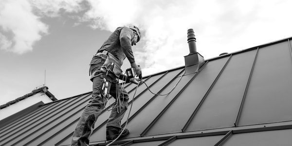 A worker in safety gear drills on a metal roof under a blue sky.
