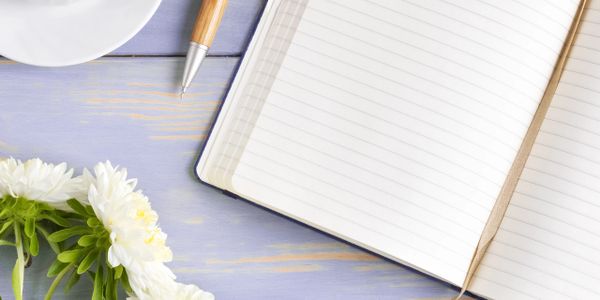 Open notebook with pen, coffee, glasses, and white flowers on a pastel wooden table.