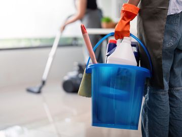 Person holding a blue cleaning bucket with supplies, another mopping the floor in the background.