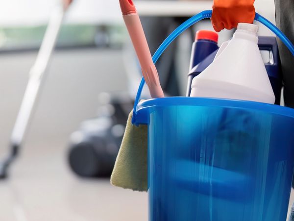 Person holding a blue cleaning bucket with supplies, another mopping the floor in the background.