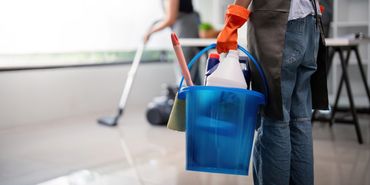 Person holding a blue cleaning bucket with supplies, another mopping the floor in the background.