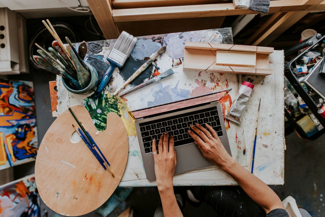 Hands typing on a laptop surrounded by art supplies.