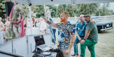 Man taking selfie with friends at outdoor DJ booth.