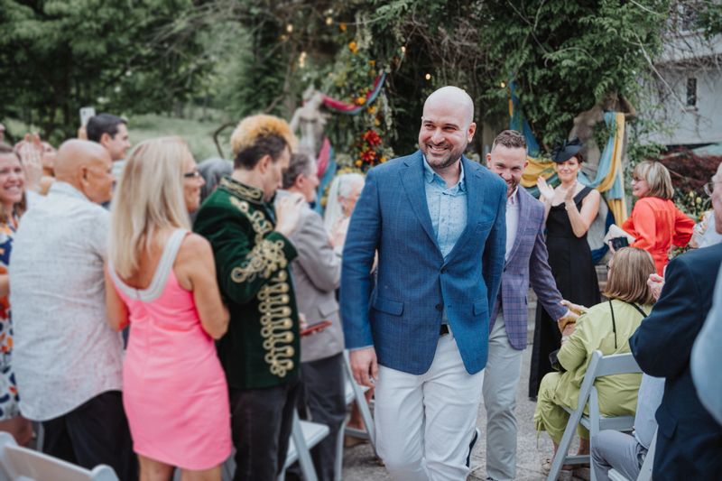 Smiling gay couple holding hands and walking through crowd of guests after their outdoor wedding ceremony.