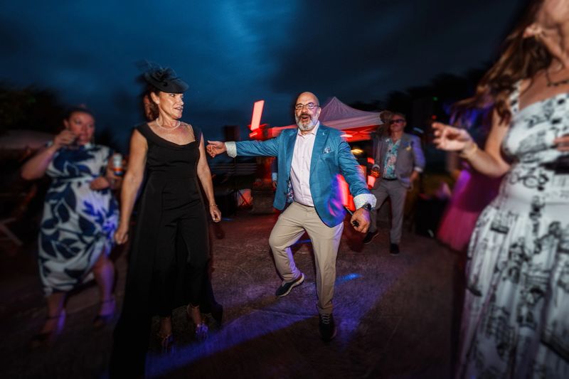 A lively and excited group of adults have fun dancing together while attending an outdoor wedding reception at night.