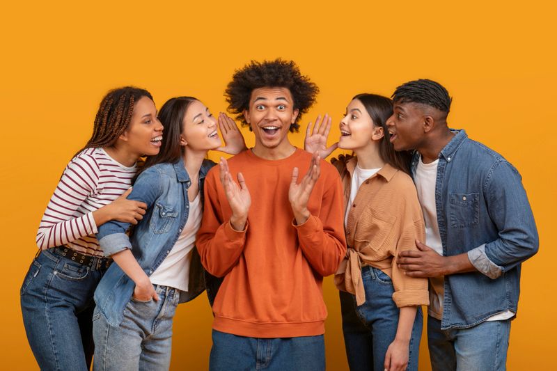 Five diverse young adults are gathered around one surprised man, sharing secrets and expressing excitement against an orange background
