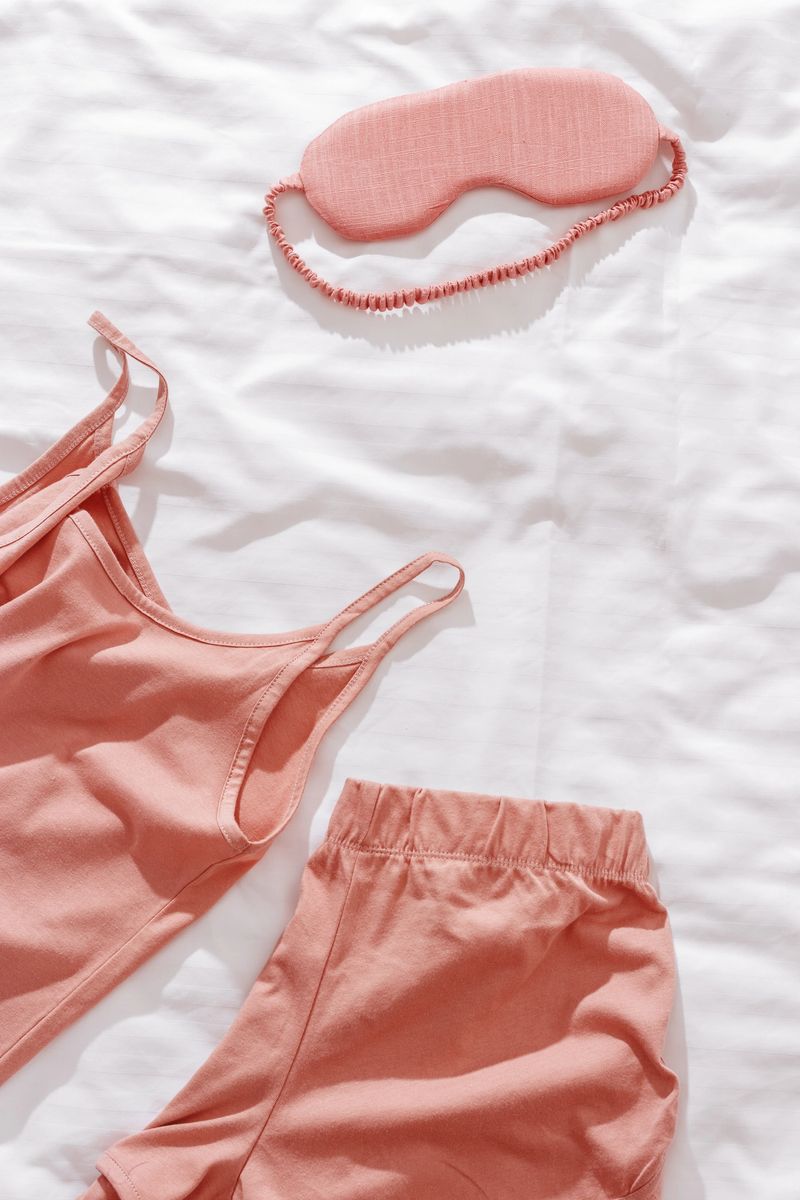 Pink female pajama and sleep eye mask on white cloth bed sheet. Top view comfort pyjamas for health sleeping. Romance mood, singlet and shorts for girl light pink trend color, sleepwear for slumber.