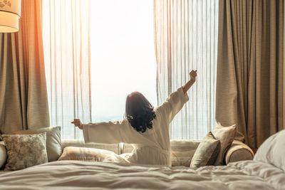 Woman's back, seen stretching in a luxurious room facing window while sitting on bed. 