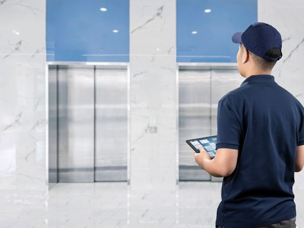Technician in blue uniform holding a tablet, inspecting elevator doors in a modern building.