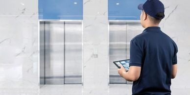 Technician in blue uniform holding a tablet, inspecting elevator doors in a modern building.