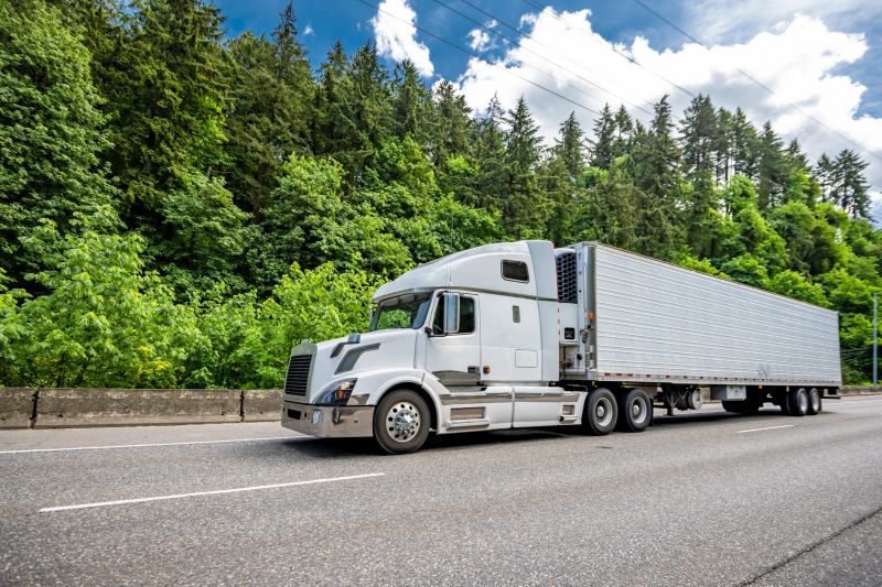 Industrial carrier white big rig semi truck tractor with extended cab for truck driver rest transporting cargo in refrigerator semi trailer running on the highway road with green forest on the side