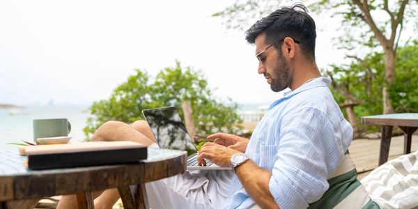 Man working on laptop outdoors by the beach, relaxing on a cushion.