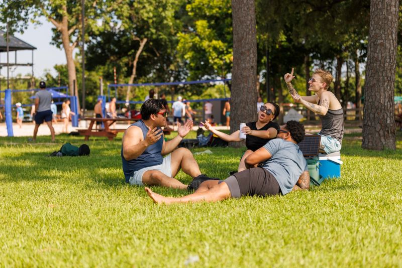 Diverse group of friends engaged in a playful card game while sitting on the green grass in a park. The scene captures two men and one woman of mixed ethnicity enjoying a casual, fun moment together outdoors. The woman, possibly Hispanic or Latino, is animatedly discussing her move, while one of the men, who appears to be Asian, listens attentively. Another man, who is African-American, seems to be sharing a light moment. The atmosphere is relaxed, with other park-goers visible in the background, indicating a communal urban park environment.