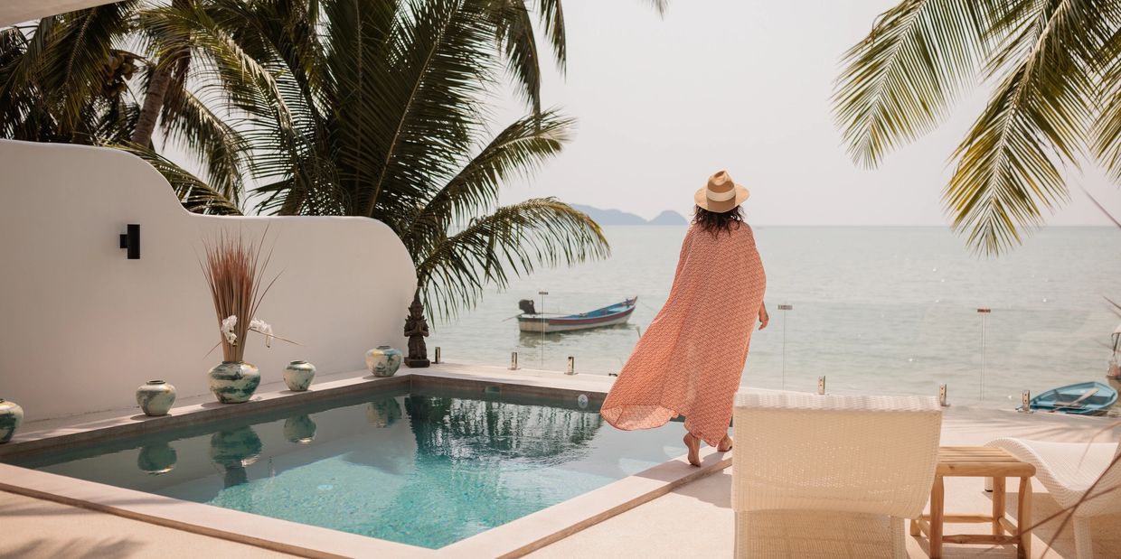 Woman in a hat stands by a pool overlooking the ocean with palm trees.