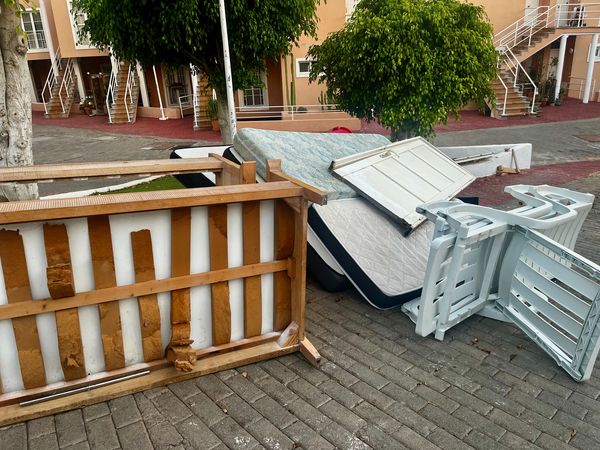 Discarded furniture including mattresses, plastic chairs, and a wooden table on a paved area.