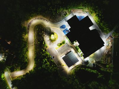 Aerial night view of a lit building and winding road surrounded by trees.