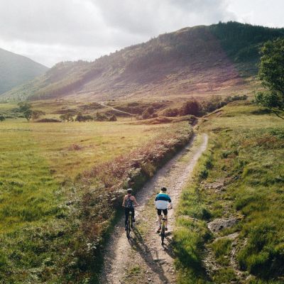 Two cyclists on a rural dirt path surrounded by green hills.