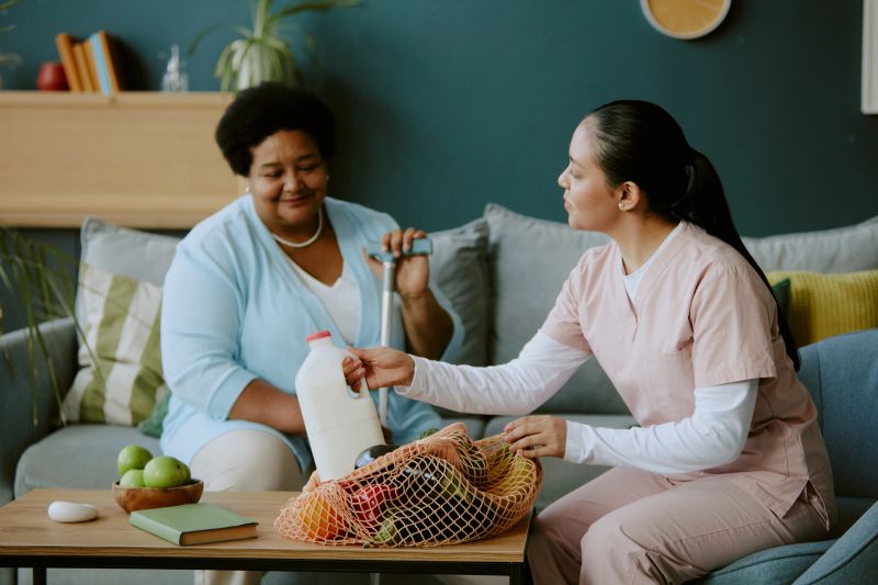 Young nurse unboxing purchase and putting them on table while senior African American female patient observing her gladly on sofa