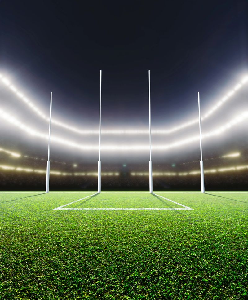 A generic seated aussie rules stadium showing goal posts on a green grass pitch at night under illuminated floodlights - 3D render