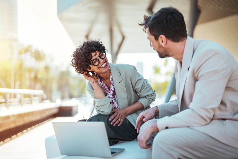 Caucasian man in a light suit and Hispanic woman with curly hair, dressed in business attire, are discussing work on a laptop while seated outside on a sunny day.