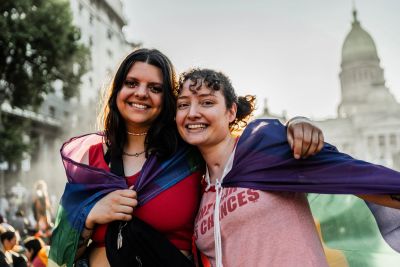 Two smiling women wrapped in a rainbow pride flag outdoors.