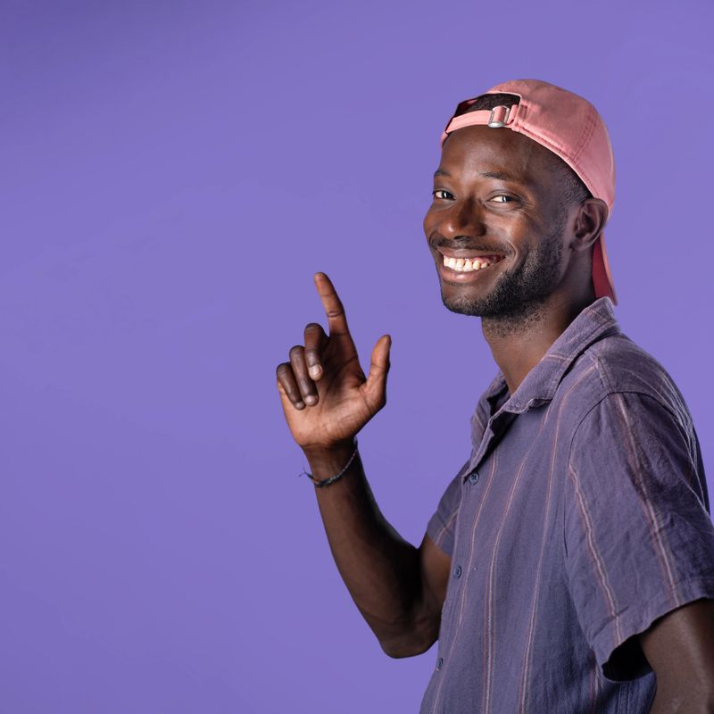 A cheerful young African American man wearing a pink cap and casual shirt, smiling and pointing up on a purple background - Happy and positive portrait - Vibrant and colorful setting.