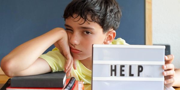 Tired boy holding a lightbox sign saying HELP at a study desk.