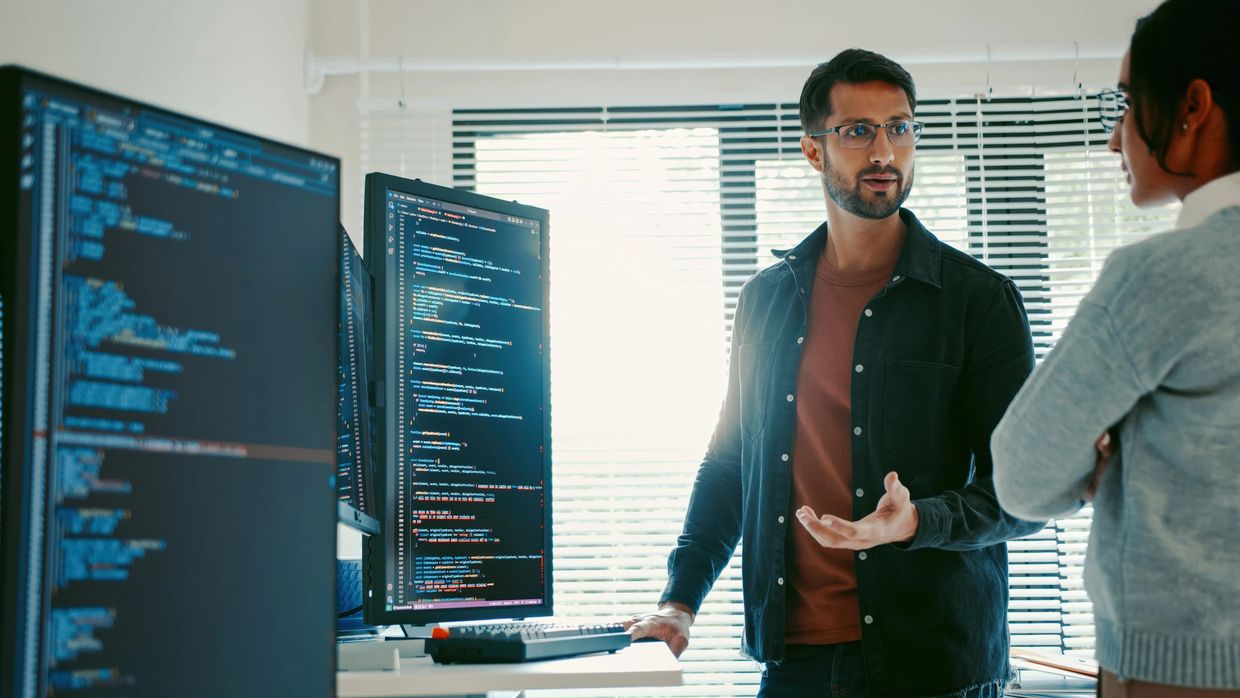 Two software developers discussing code displayed on dual monitors in a modern office.
