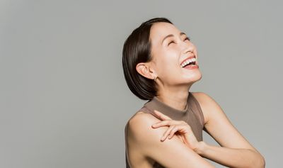 Woman laughing with her head tilted back, arms crossed, sleeveless top, and a neutral background.