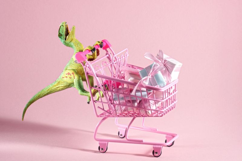 Funny green dinosaur toy with shopping cart full of present boxes on a soft pink background.