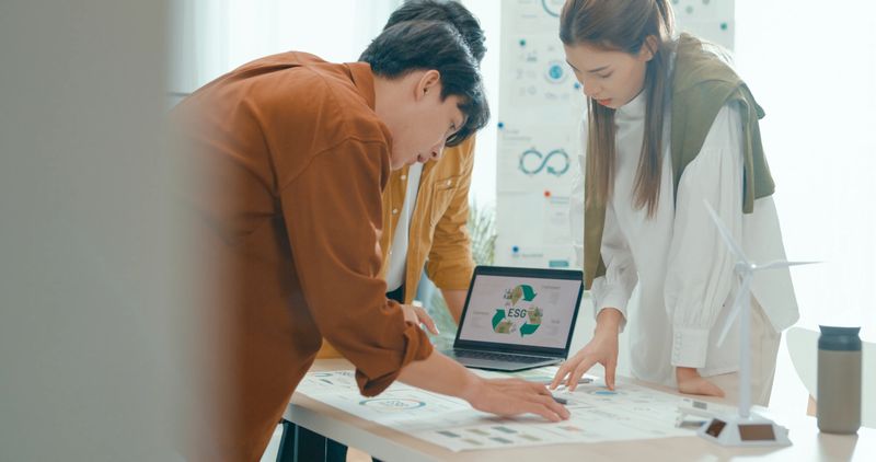 Team members examine ESG-related documents, pointing and discussing investment strategies for sustainable and renewable energy resources. Eco-Friendly Business Team Discussing ESG Goals concept.