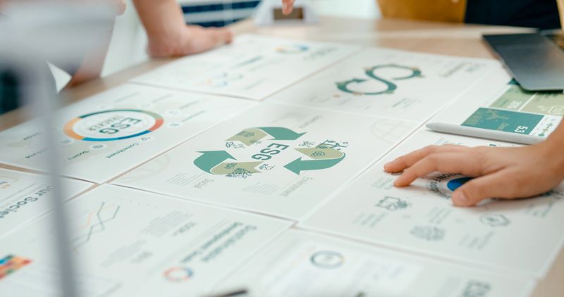 Closeup of team members examine ESG-related documents, pointing and discussing investment strategies for sustainable and renewable energy resources. Eco-Friendly Business Discussing ESG Goals concept.