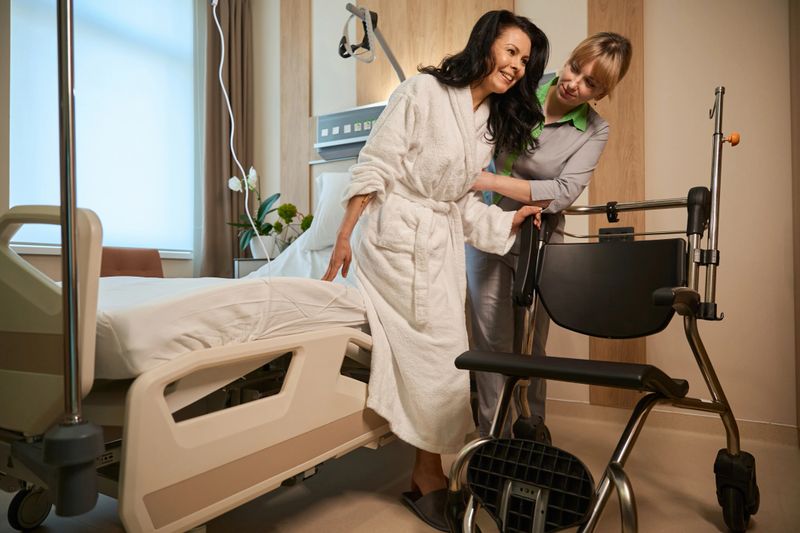 Caring nurse helps a clinic patient transfer from bed to wheelchair, the ward has modern equipment