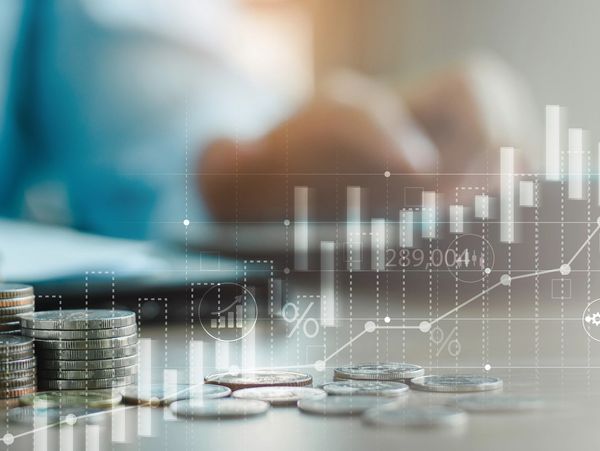 Stacks of coins with financial graphs overlay and person writing in background.