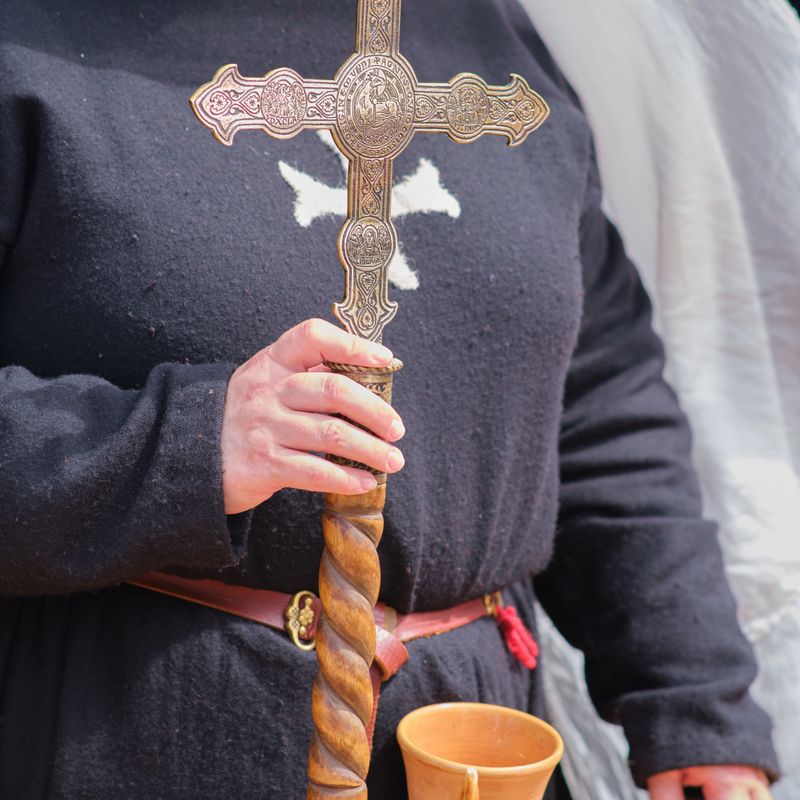 A person dressed in medieval attire holds a detailed cross with intricate engravings in one hand and a wooden cup in the other. They wear a black garment with a white cross on the chest