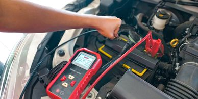 A person testing a car battery with a red battery tester under the hood.
