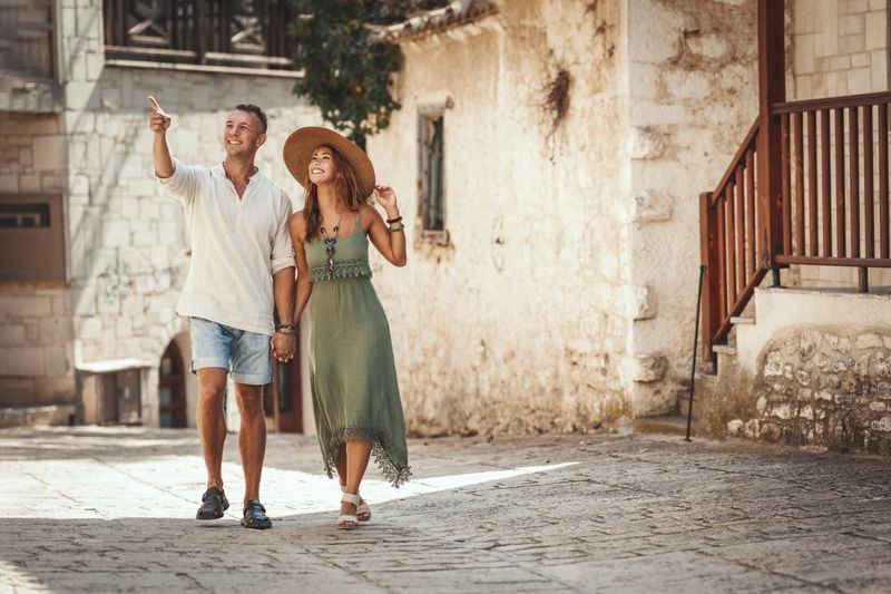 A beautiful young couple is having fun while walking around a Mediterranean town. They are enjoyed in summer sunny day, holding each other's hand and smiling.